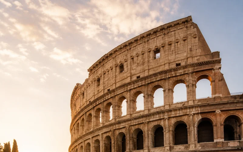 A photorealistic 16:9 wide-angle scene of the Roman Colosseum at golden hour, shot from street level with warm evening light across the ancient stone arches and a quiet foreground cobblestone street. Alt: Day-by-day Italy itinerary starting point at the Colosseum in Rome.